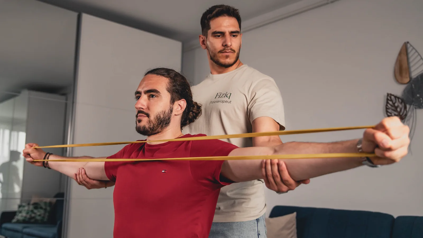 Fizioterapeut Fiziq care asistă un pacient în timpul unui exercițiu cu bandă elastică pentru mobilitatea și stabilitatea umerilor, într-o ședință de recuperare la domiciliu.